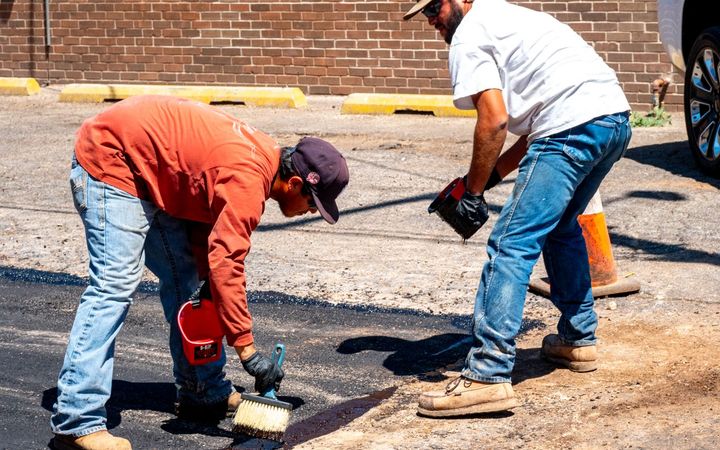 Two workers patching asphalt beside a brick building